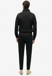 Man in een volledig zwarte casual outfit met een lange mouw top en broek, met de rug naar de kijker, gecombineerd met lichtbeige hoge sneakers.