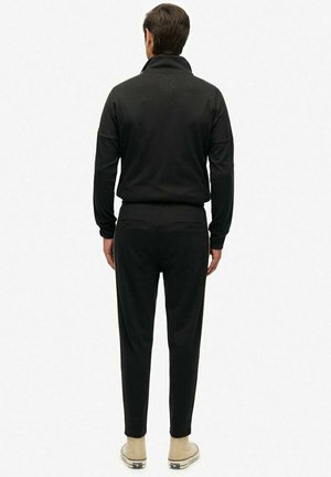 Man in een volledig zwarte casual outfit met een lange mouw top en broek, met de rug naar de kijker, gecombineerd met lichtbeige hoge sneakers.