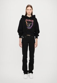 Felpa nera corta con stampa grafica, abbinata a jeans neri aderenti e sneakers bianche. Presenta un cappuccio con cordino e maniche a sbuffo.