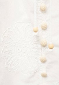 Bílý látkový top sekvětinovou výšivkou, dekorativními tkanými knoflíky a dřevěnými korálky podél předního zapínání. Jemná textura a designové detaily.