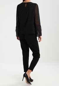 Blusa nera trasparente con motivo a pois argentati, maniche lunghe con dettagli a fiocco; abbinata a pantaloni neri a sigaretta e scarpe con tacco nere.