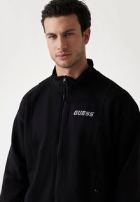 Svart zip-up jacka i mjukt material. Har hög krage och en silverprägling av "GUESS"-logotypen på övre vänstra sidan.