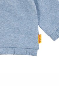 Sudadera azul clara con mangas acampanadas y dobladillo. Presenta una etiqueta amarilla con texto rojo en el lateral. Tejido suave y con textura.
