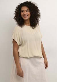 Light beige, short-sleeve blouse with a flowing design and round neckline, paired with a textured, light-colored skirt.