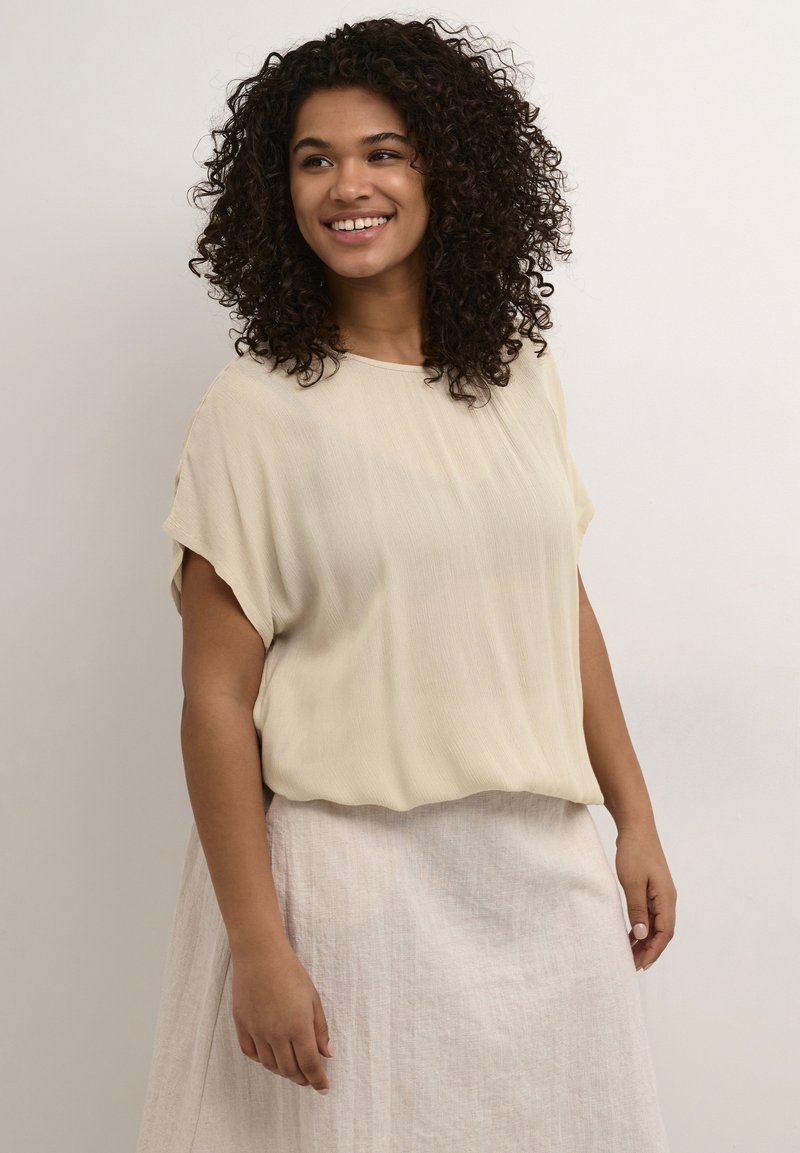 Light beige, short-sleeve blouse with a flowing design and round neckline, paired with a textured, light-colored skirt.