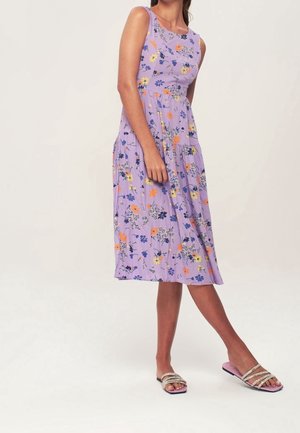 Mouwloze lavendelkleurige midijurk met oranje, gele en blauwe bloemenprint, gedragen met roze platte sandalen met riempjes met strass-steentjes.