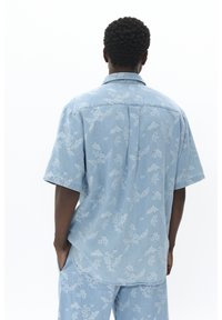 Camicia di jeans azzurro chiaro a maniche corte con un sottile motivo floreale, taglio dritto e dettagli sulla parte posteriore.