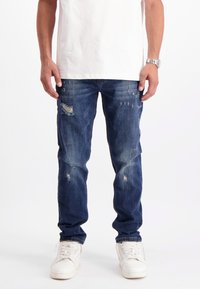 Man in versleten blauwe jeans, witte sneakers, een wit T-shirt en een zilveren polshorloge, staand tegen een witte achtergrond.