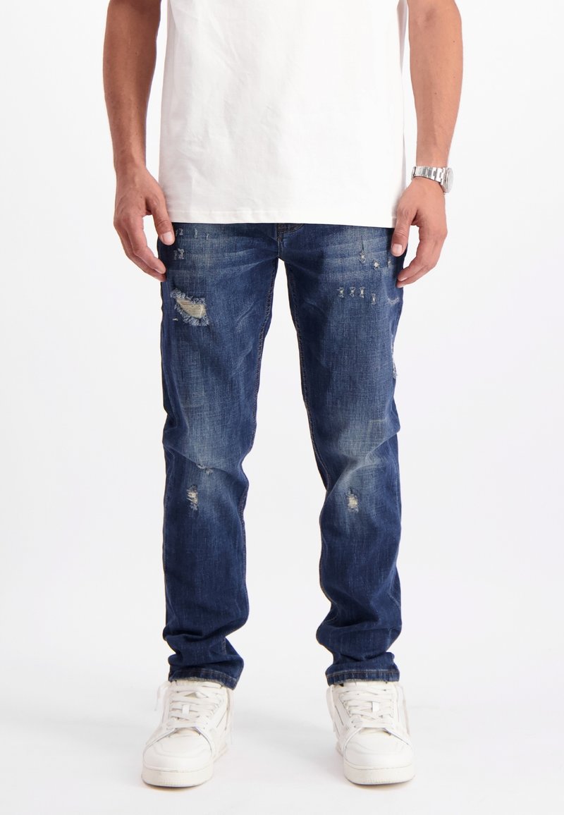 Man in versleten blauwe jeans, witte sneakers, een wit T-shirt en een zilveren polshorloge, staand tegen een witte achtergrond.