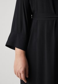 Vêtement noir à coupe décontractée, avec une taille ceinturée et des manches larges. Texture douce avec une finition lisse, mettant en avant la simplicité du design.