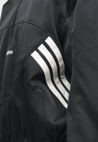 Μαύρο μπουφάν Adidas με λευκό σχέδιο τριών ρίγων στο μανίκι και μερικά ορατό λογότυπο στο στήθος.