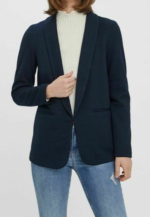 Femme en blazer texturé bleu marine foncé sur un haut blanc côtelé et un jean bleu clair déchiré, posant devant un fond clair uni.