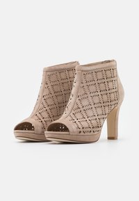 Botas de tobillo de ante beige con patrones florales cortados a láser, con punta descubierta y tacones anchos sobre un fondo blanco.
