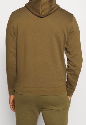 Man in olijfgroene hoodie en bijpassende trainingsbroek, handen ontspannen langs het lichaam, gezien van achteren tegen een effen achtergrond.