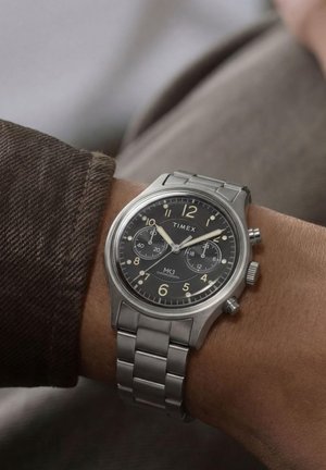 Ρολόι χειρός Timex MK1 με χρονογράφο, μαύρο καντράν, ασημένια κάσα και μεταλλικό μπρασελέ σε καρπό ατόμου που δείχνει 10:10:38.