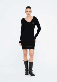 Sort strikket kjole med V-hals, lange ærmer og kontrasterende hvide kanter ved ærmekanter og underkant. Modellen bærer sorte knæhøje støvler med spænder.