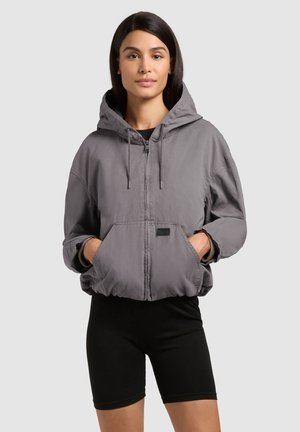 Kobieta ubrana w szarą, zapinaną na zamek bluzę z kapturem, z rękami w przednich kieszeniach, oraz czarne spodenki rowerowe na jednolitym tle.