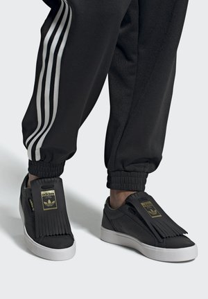 Sneaker nere con finitura in pelle liscia, dotate di un dettaglio frangiato sulla linguetta e suola in gomma bianca. Accenti del logo dorato visibili.