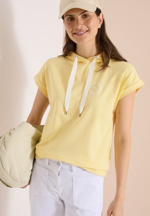Donna che indossa una felpa gialla a maniche corte con coulisse bianche, pantaloni bianchi, cappello beige, tiene una giacca imbottita beige, sorridente su uno sfondo neutro.