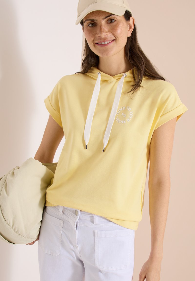 Femme portant un sweat à capuche jaune à manches courtes avec des cordons blancs, pantalon blanc, casquette beige, tenant une doudoune beige, souriante devant un fond neutre.