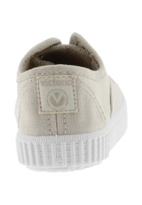 Beige lærredssneaker med en tekstureret overdel, hvid gummisål og et logomærke på bagsiden, med et forenklet design uden synlige mønstre.