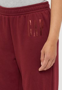 Pantaloni della tuta color marsala in morbido tessuto con cintura elastica. Presentano un logo "GAP" rosa oro lucido sul lato. Design semplice.