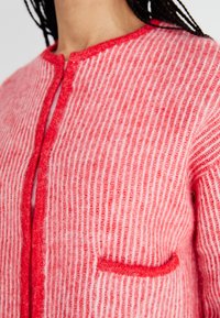 Adīta kardigāna no mīkstas rozā vilnas ar sarkanu svītru rakstu, apaļa izgriezuma, un neliela kabatiņa kreisajā pusē.