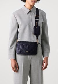 Prošívaná kabelka přes rameno v námořnické modři z kůže s pozlacenými detaily, zipovým uzávěrem a odnímatelným držákem na kartu, s logem na přední straně.