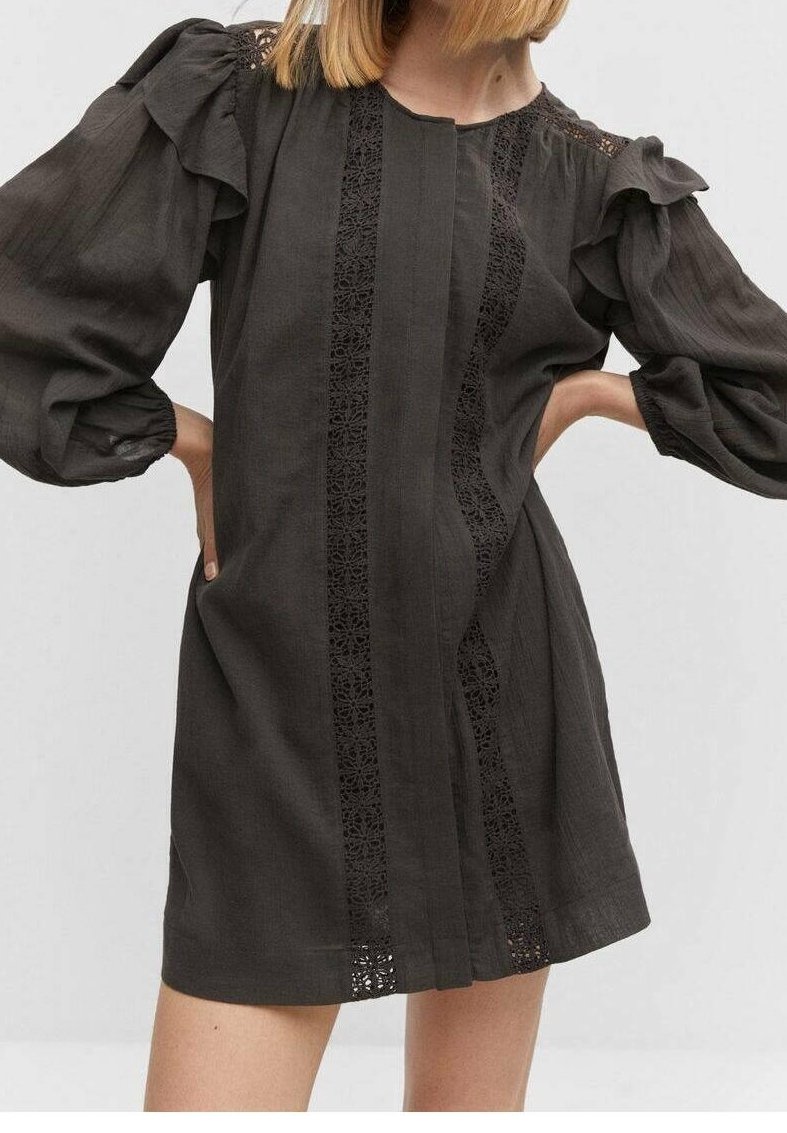 Femme portant une robe noire à manches longues avec des manches bouffantes et un détail en dentelle verticale à l'avant, les mains posées sur les hanches.