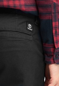 Pantaloni neri con una texture liscia, dotati di un logo Timberland sulla tasca posteriore, abbinati a una camicia a quadri rossa e blu.