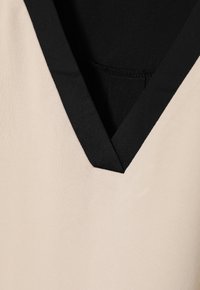 Béžový top s hlbokým výstrihom do V a kontrastným čiernym lemovaním, vyrobený zo hladkej látky. Dizajn je jednoduchý a minimalistický.