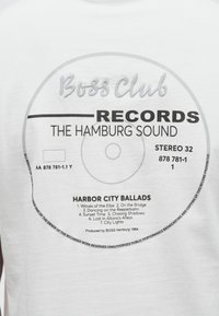 Valge t-särk, millel on plaatide kujundus musta tekstiga. Loetletud laulud rubriigi "Harbor City Ballads" all, graafikaga, mis meenutab vinüülplaadi etiketti.