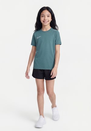 Fille marchant en short noir, t-shirt Nike bleu sarcelle et chaussures de sport blanches sur un fond blanc uni.