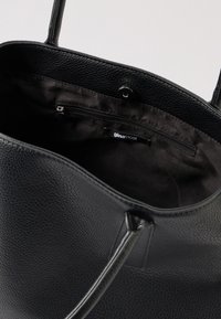 Crna torba od teksturirane kože s unutarnjim zip džepom i zatvaračem na klik, prikazuje "ginatricot" oznaku branda unutra.