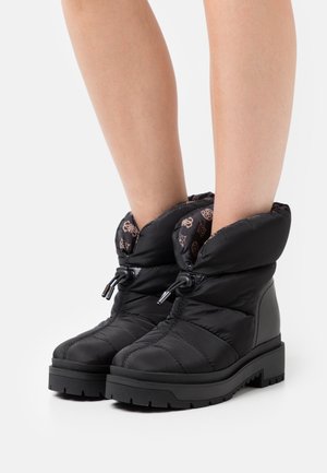 Botas negras acolchadas hasta el tobillo con suelas gruesas y cierres de cordones ajustables, llevadas sobre piernas desnudas.