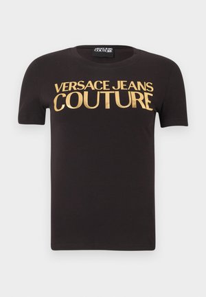 Črna majica s kratkimi rokavi z napisom "Versace Jeans Couture" v velikih zlatih črkah čez prsa, okrogel izrez in notranja etiketa iz blaga.