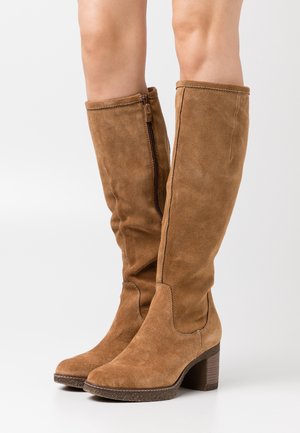 Bottes en daim marron hauteur genou avec talons blocs et fermetures éclair latérales, portées sur des jambes nues sur fond blanc.