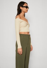 Cropped top beige a coste con design off-shoulder e maniche lunghe, abbinato a pantaloni a vita alta verde oliva con zip.