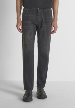 Man met zwarte, verweerde, rechte jeans en zwarte leren laarzen tegen een effen grijze achtergrond.