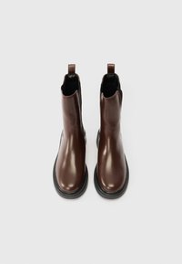 Pár lesklých hnědých kožených kotníkových bot s černými elastickými stránkami a poutky, zobrazených z výšky na bílém pozadí.