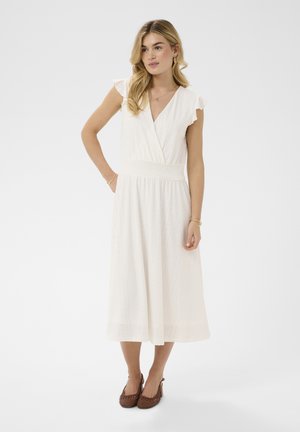 Žena s dlhými blond vlasmi, ktorá má na sebe biele šaty midi s očkami a krátkymi rukávmi, a hnedé pletené podpätky, stojaca na jednoduchom pozadí.