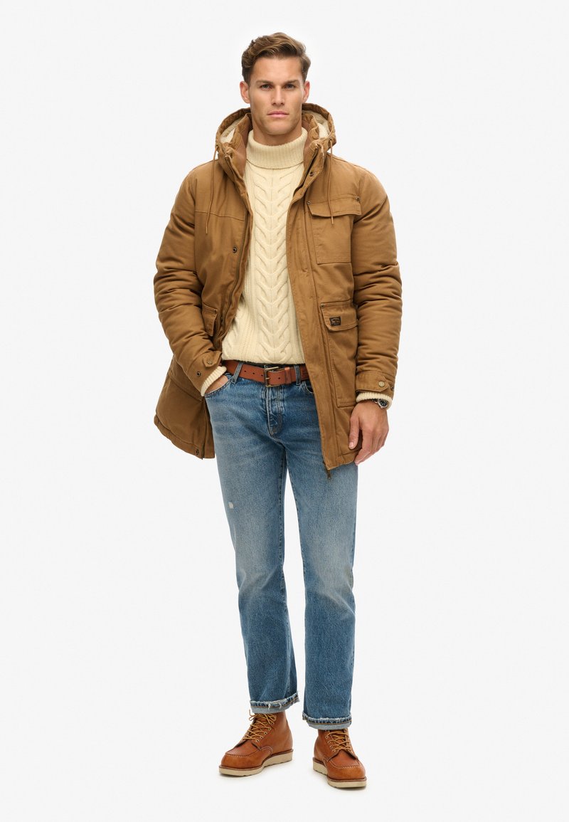 Bruine gewatteerde jas met capuchon, crèmekleurige kabelgebreide trui, blauwe spijkerbroek en bruine veterlaarzen. De jas heeft voorzakken en een rits.