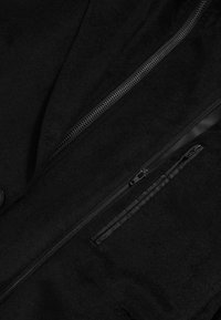 Sort uldjakke med lynlås, to lynlåsede lommer og en glat tekstur. Bemærkelsesværdigt hårdvarer inkluderer en knap og lynlåse.