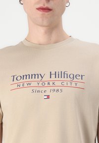 Beige bomull t-skjorte med rund hals. Fronten har "Tommy Hilfiger New York City" i marineblå og rød skrift, med logo-aksent.