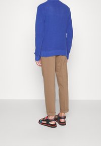 Pull en maille bleu, pantalons beiges, sandales noires avec des brides et des accents colorés, vues de dos sur fond blanc.