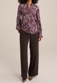 Woman wearing a patterned purple blouse and dark brown wide-leg trousers, paired with pointed-toe taupe heels, standing against a plain background.