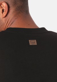 Černý svetr s žebrovaným výstřihem, na zádech je obdélníková hnědá značková etiketa s písmeny "LVM."