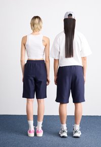 Deux personnes se tiennent de dos, portant des hauts blancs et des shorts marins. La personne à gauche a un tatouage, et les deux portent des chaussures de sport de couleurs distinctes.