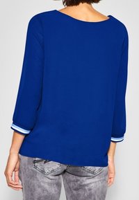 Blå, löst sittande topp med trekvartslånga ärmar, med vita randiga detaljer vid muddarna. Slät tyg, rundad halsringning.