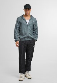 Sportovní sako v matné smaragdové barvě se zipem vpředu, ribbovanými manžetami a logem. Spárováno s černými jogger kraťasy na elastický pas a světlými teniskami.